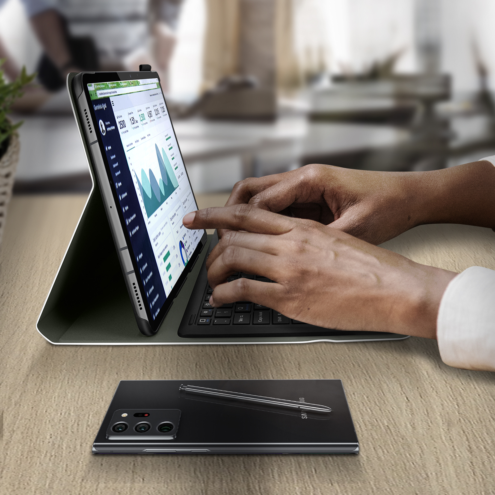 Eine Person tippt auf einem Tablet mit Tastatur. Auf dem Tisch liegen ein Telefon und ein Stift. Auf dem Bildschirm sind Diagramme zu sehen.