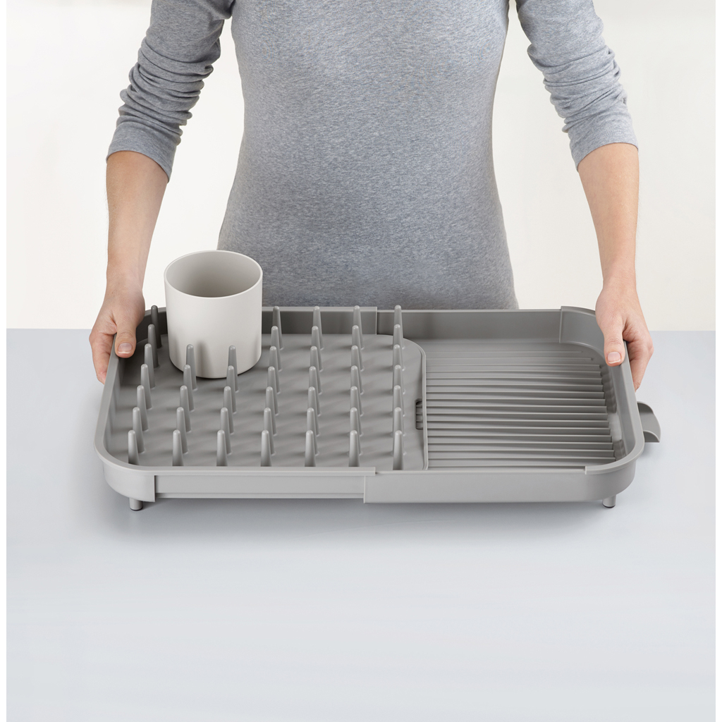 Una persona sostiene un escurridor gris con una taza blanca. El escurridor tiene una sección con clavijas y un área acanalada.