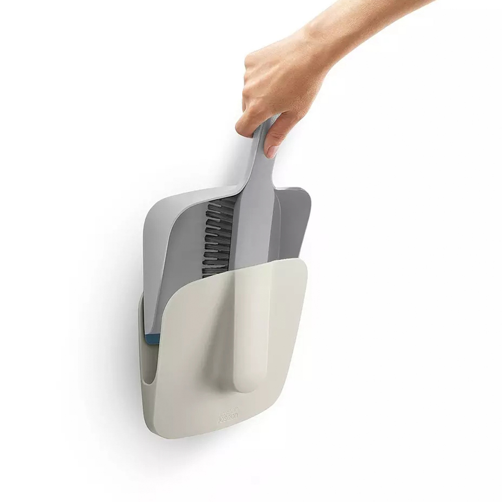Una mano sostiene un juego de cepillo y recogedor gris. El juego está colgado en una pared blanca. El juego es gris y blanco.