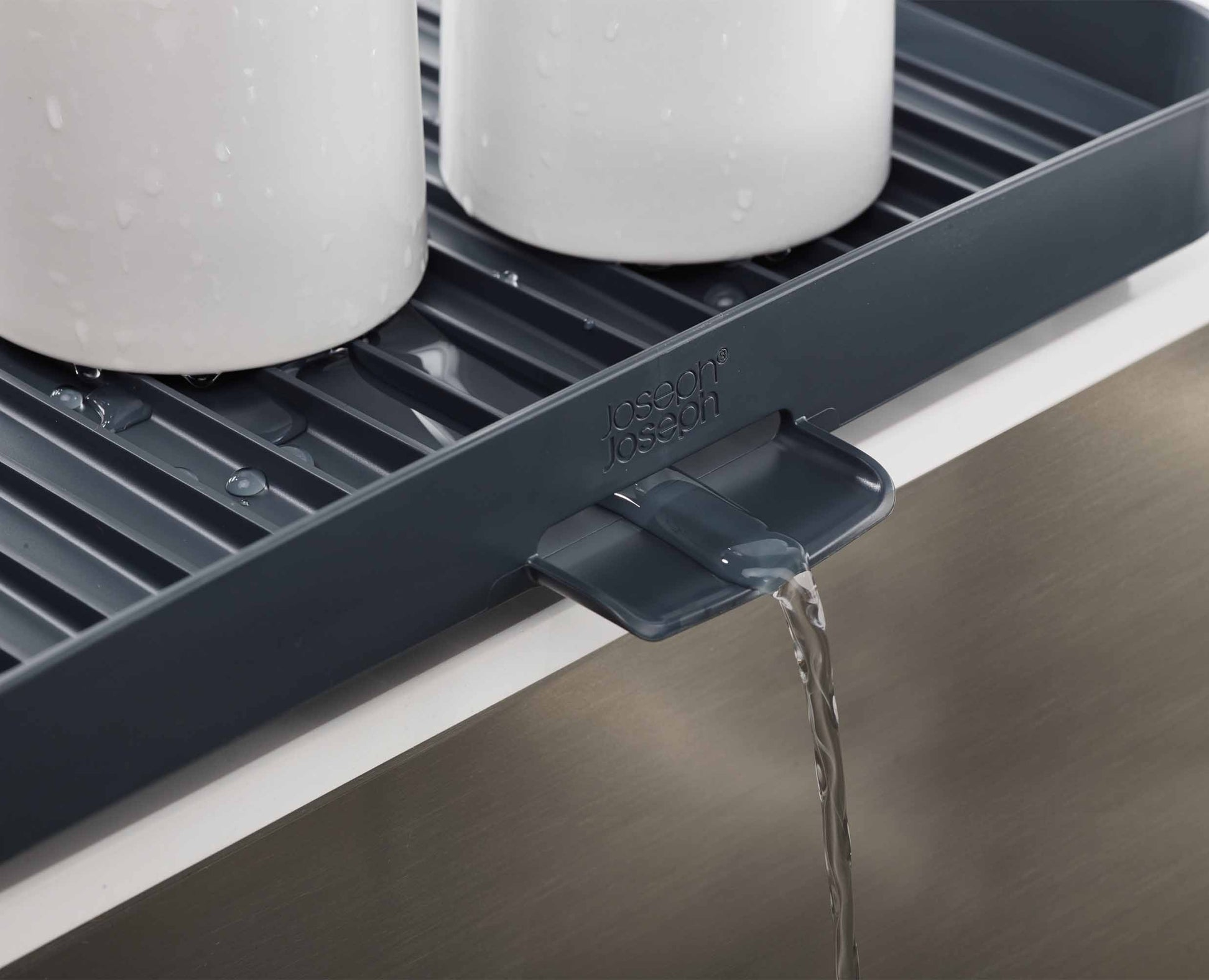 Escurridor de platos gris oscuro con dos tazas blancas. El agua drena de la boquilla a un fregadero de metal.