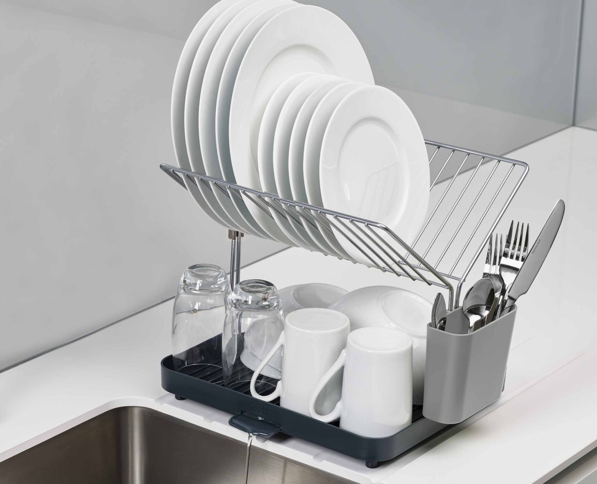 Un escurreplatos con platos, vasos, tazas y cubiertos. El estante está encima de un fregadero. Platos, tazas y tazas blancos.