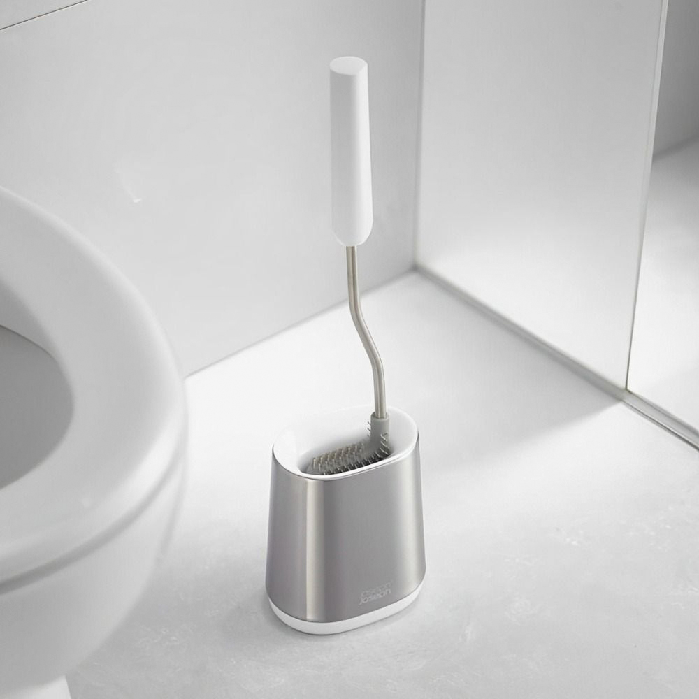 Un cepillo de inodoro en un soporte se encuentra cerca de un inodoro. El cepillo es blanco y plateado. El soporte es plateado y blanco.