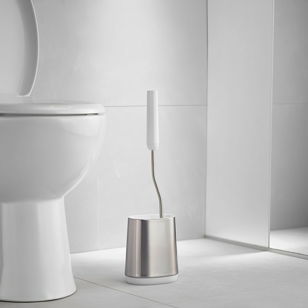 Un inodoro blanco y un soporte de cepillo de metal en un baño con azulejos blancos. La tapa del inodoro está levantada.