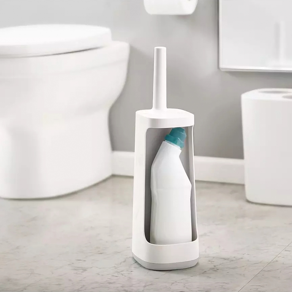 Un soporte blanco para cepillo de inodoro con una botella de producto de limpieza dentro. Un inodoro, rollo de papel y cubo de basura en el fondo.