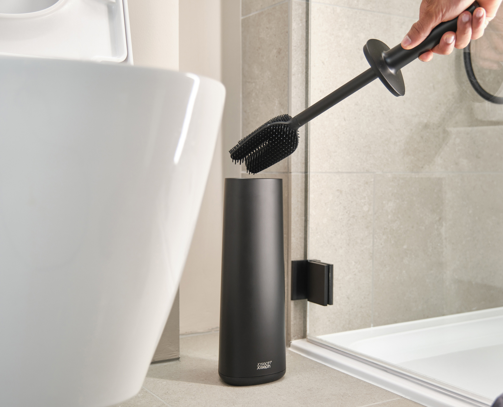 Un cepillo de inodoro negro sostenido sobre un recipiente negro en un baño. Un inodoro blanco está a la izquierda.