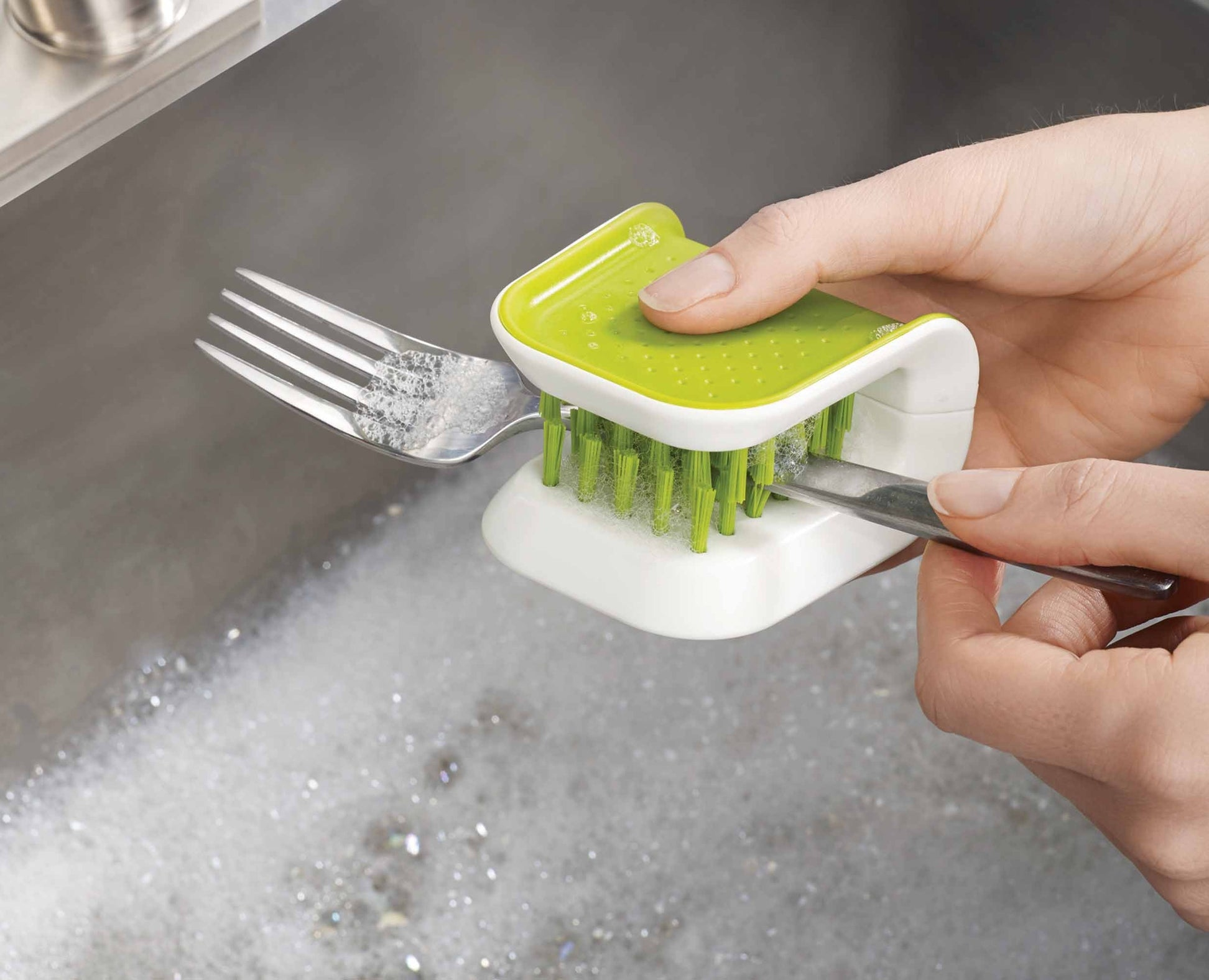 Una persona lava una forchetta e un coltello con una spazzola verde e bianca in un lavandino pieno di acqua saponata.