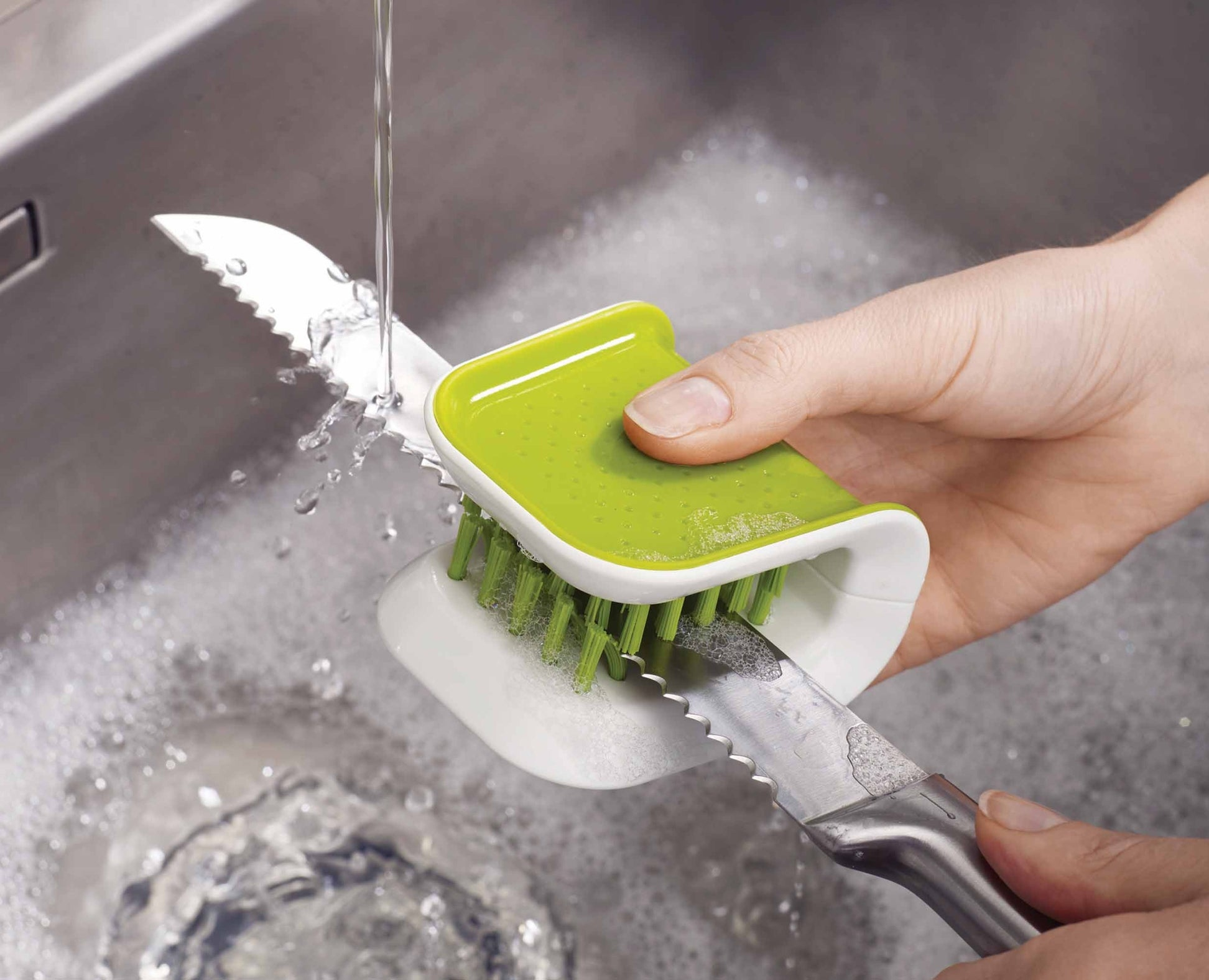 Una mano lava un coltello con una spazzola verde in un lavandino pieno di acqua saponata. L'acqua scorre sul coltello.