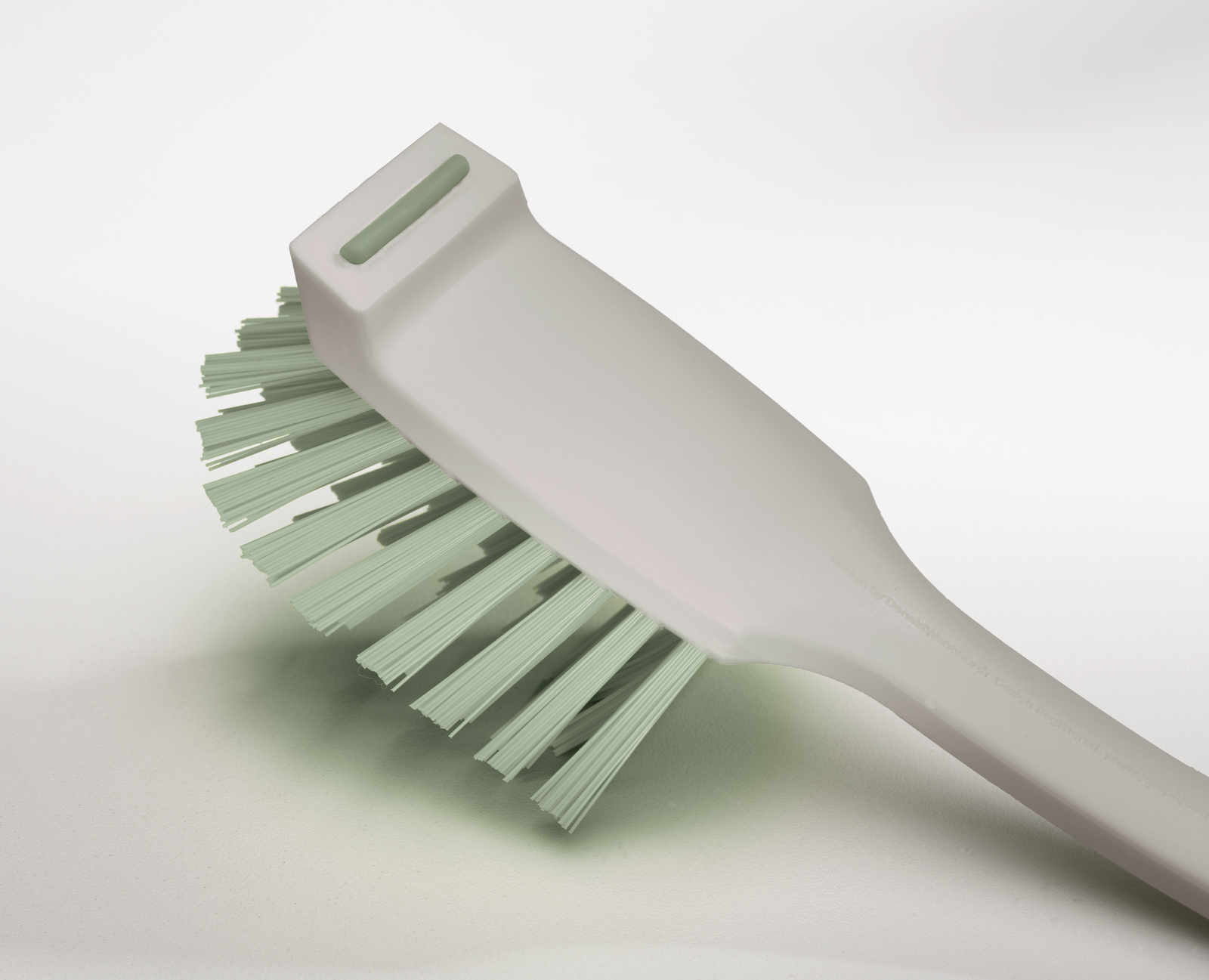 Primer plano de un cepillo de limpieza gris claro con cerdas verdes sobre un fondo blanco. El cepillo está en ángulo.