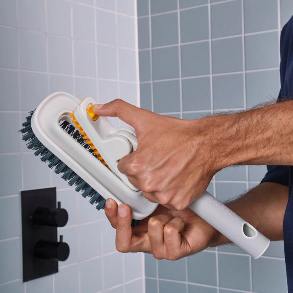 Una persona sostiene un cepillo de limpieza en un baño. El cepillo es blanco con cerdas grises y detalles amarillos. El fondo es de baldosas.