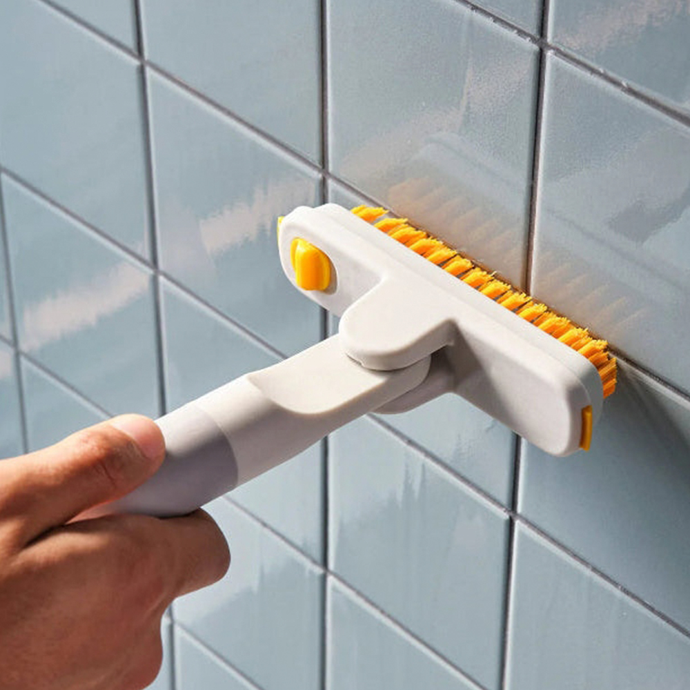 Mano sosteniendo un cepillo blanco y amarillo contra una pared de azulejos azules. El cabezal del cepillo tiene cerdas naranjas, utilizado para limpiar.