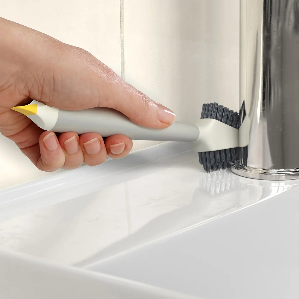 Mano sosteniendo un cepillo de limpieza gris y blanco con punta amarilla, frotando alrededor de un grifo cromado. Fregadero y azulejos blancos en el fondo.