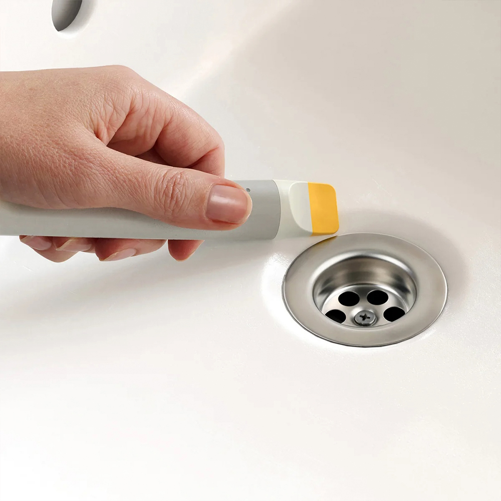 Mano sosteniendo una herramienta de limpieza con cabeza amarilla, cerca de un desagüe de lavabo. El lavabo y la herramienta son blancos.