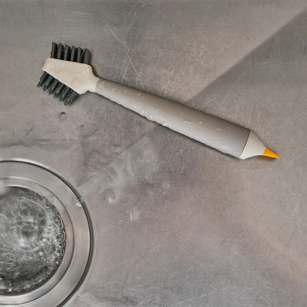 Un cepillo de limpieza gris y blanco yace sobre una superficie húmeda de acero inoxidable, cerca de un desagüe. El cepillo tiene cerdas oscuras.