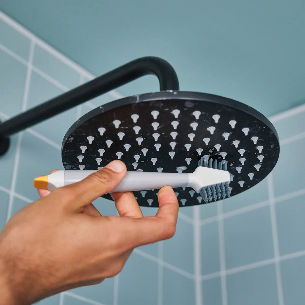 Una mano limpia un cabezal de ducha negro con un cepillo gris y amarillo. El fondo es una pared de azulejos azules y el brazo de la ducha.