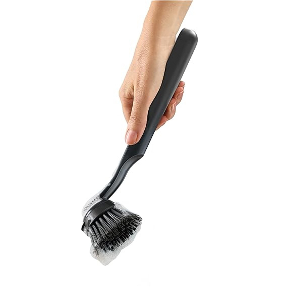 Una mano sostiene un cepillo negro para lavar platos con cerdas jabonosas. El cepillo está inclinado hacia abajo, con espuma en la base.