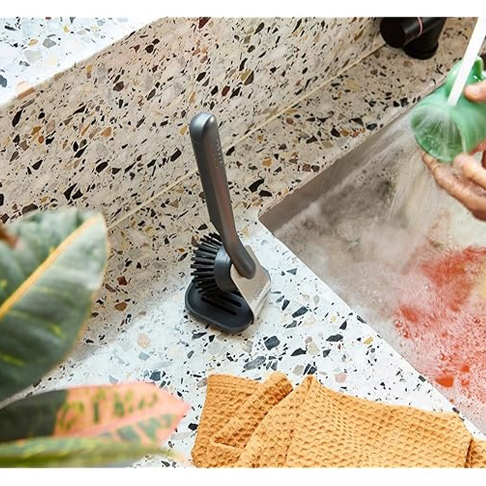 Un cepillo gris para platos está sobre una encimera moteada. Se está lavando una taza verde. Un paño naranja y hojas de plantas están en primer plano.