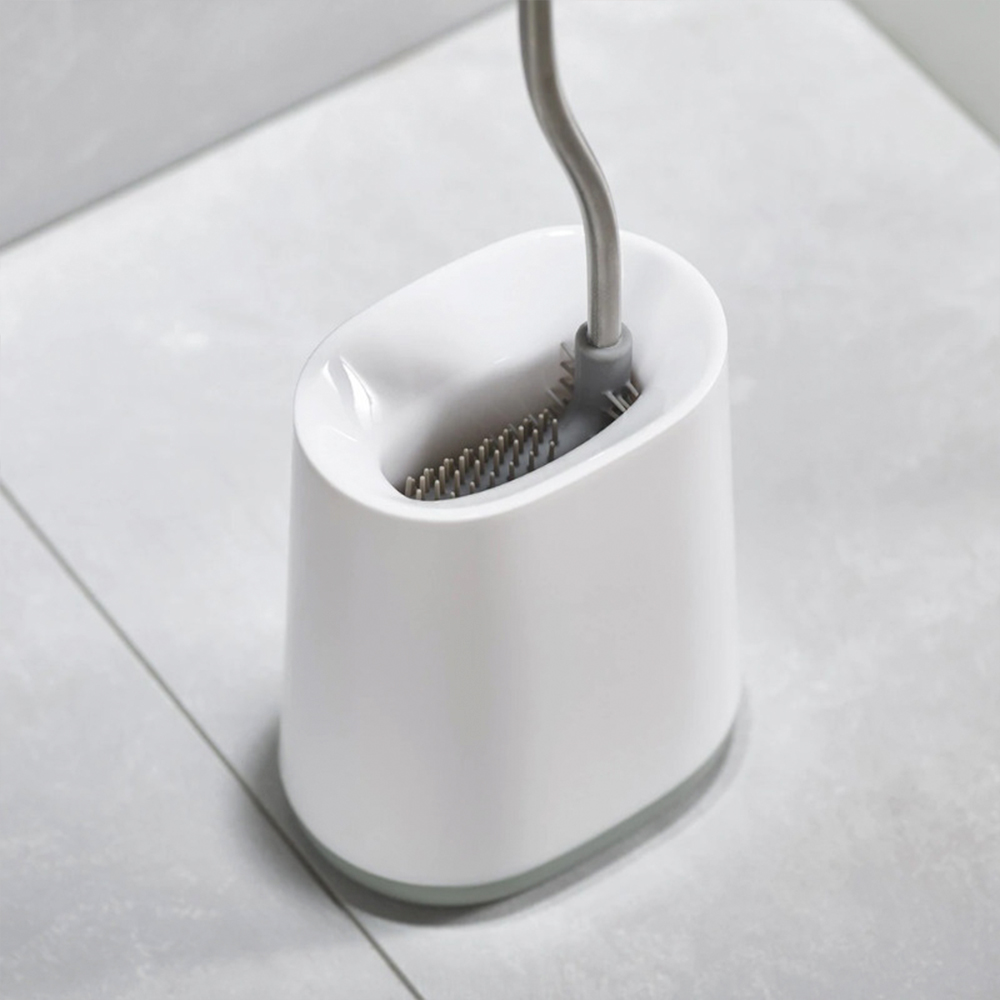 Un soporte blanco para cepillo de inodoro con base gris y cepillo gris. El fondo es un suelo de baldosas blancas.