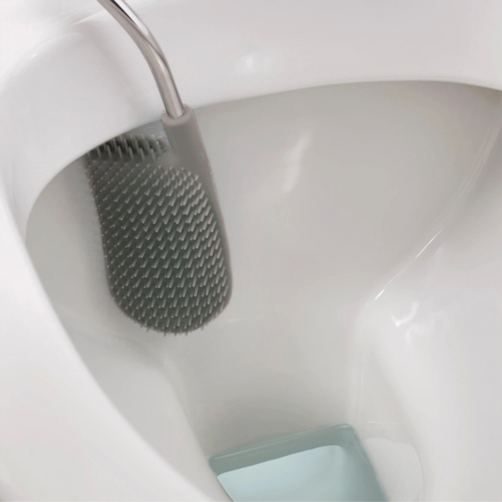 Un cepillo de inodoro gris está dentro de un inodoro blanco. El cabezal del cepillo tiene textura. El inodoro está limpio, con fondo azul-verde.