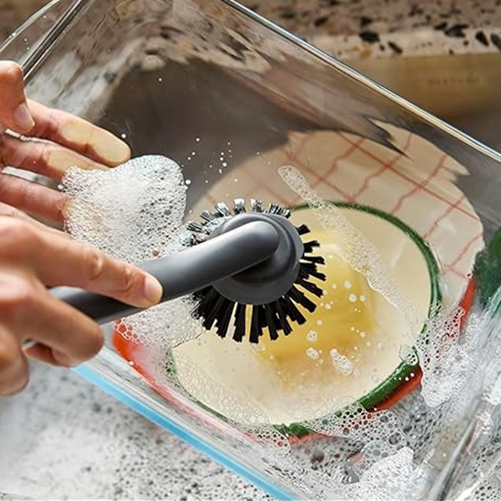 Una persona lava los platos. Un cepillo gris friega un plato en un recipiente transparente con agua jabonosa. Es visible un paño de cocina.