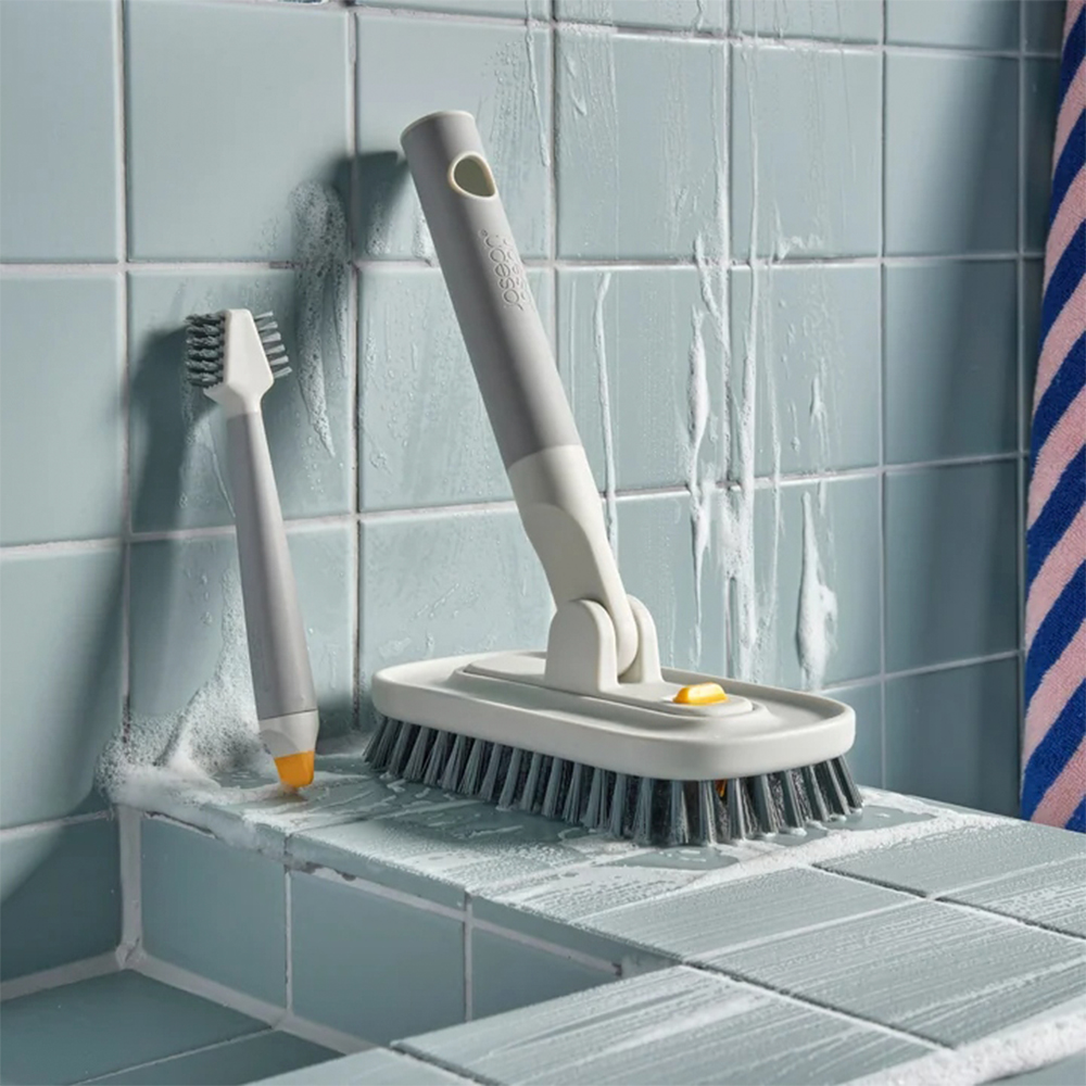 Herramientas de limpieza de baño: un cepillo de mango largo y un cepillo más pequeño. Ambos son grises y blancos con cerdas oscuras, sobre baldosas azules con jabón.