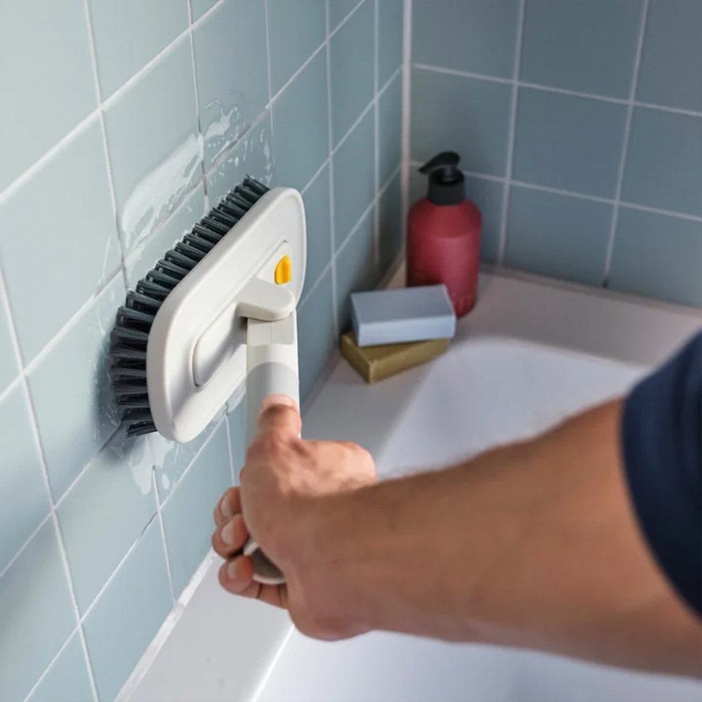 Una persona limpia la pared del baño con un cepillo. Se ven jabón y una botella. La pared tiene baldosas azules.