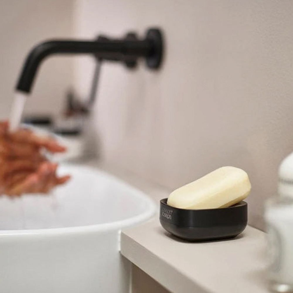 Escena de baño. Grifo negro, lavabo blanco. Jabón en plato negro. Una mano se lava.