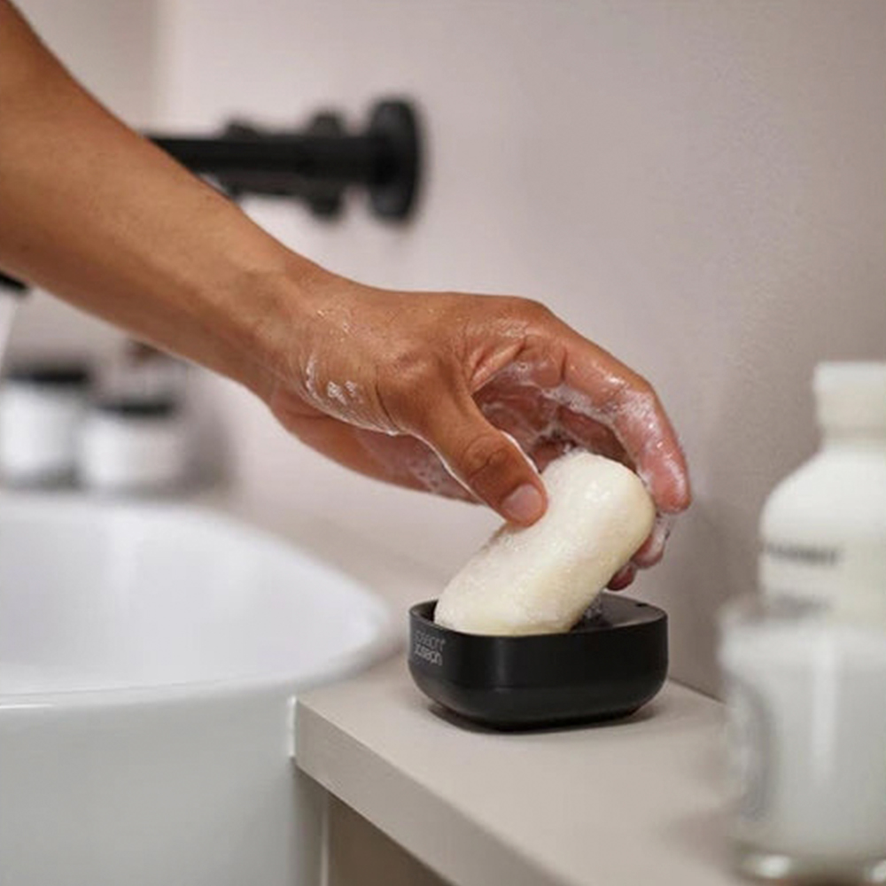 Una mano con espuma de jabón alcanza una pastilla de jabón en un jabonera negra sobre una encimera de baño. Un lavabo está en el fondo.