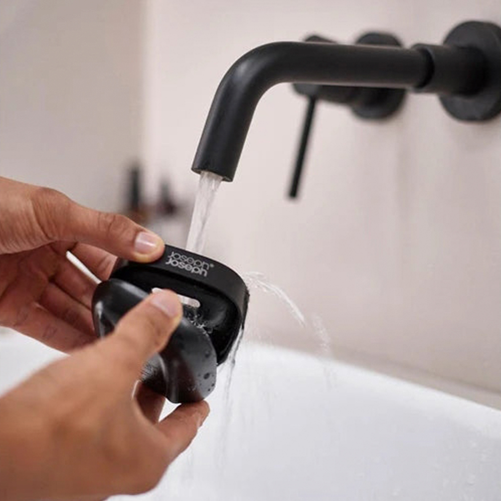 Manos sosteniendo un objeto negro bajo un grifo con agua corriendo. El grifo es negro y está montado en una pared blanca.