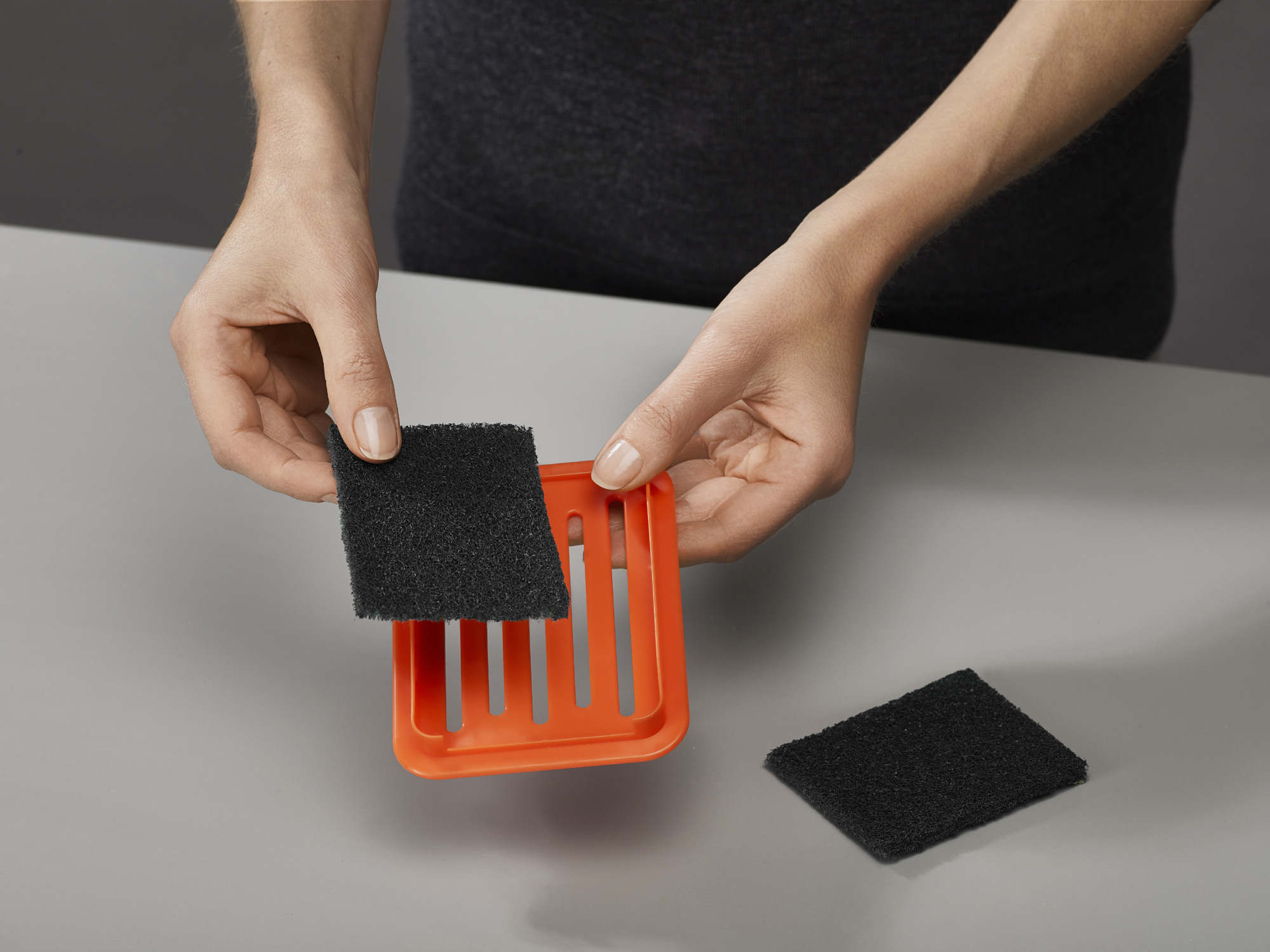 Manos sostienen un cuadrado de filtro negro sobre un marco naranja. Otro filtro yace sobre la superficie gris. Camisa oscura al fondo.