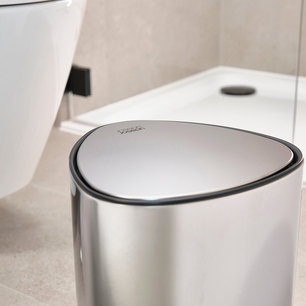 Un cestino metallico con coperchio triangolare è a fuoco. Un WC bianco e un piatto doccia sono sullo sfondo. La scena è in un bagno.