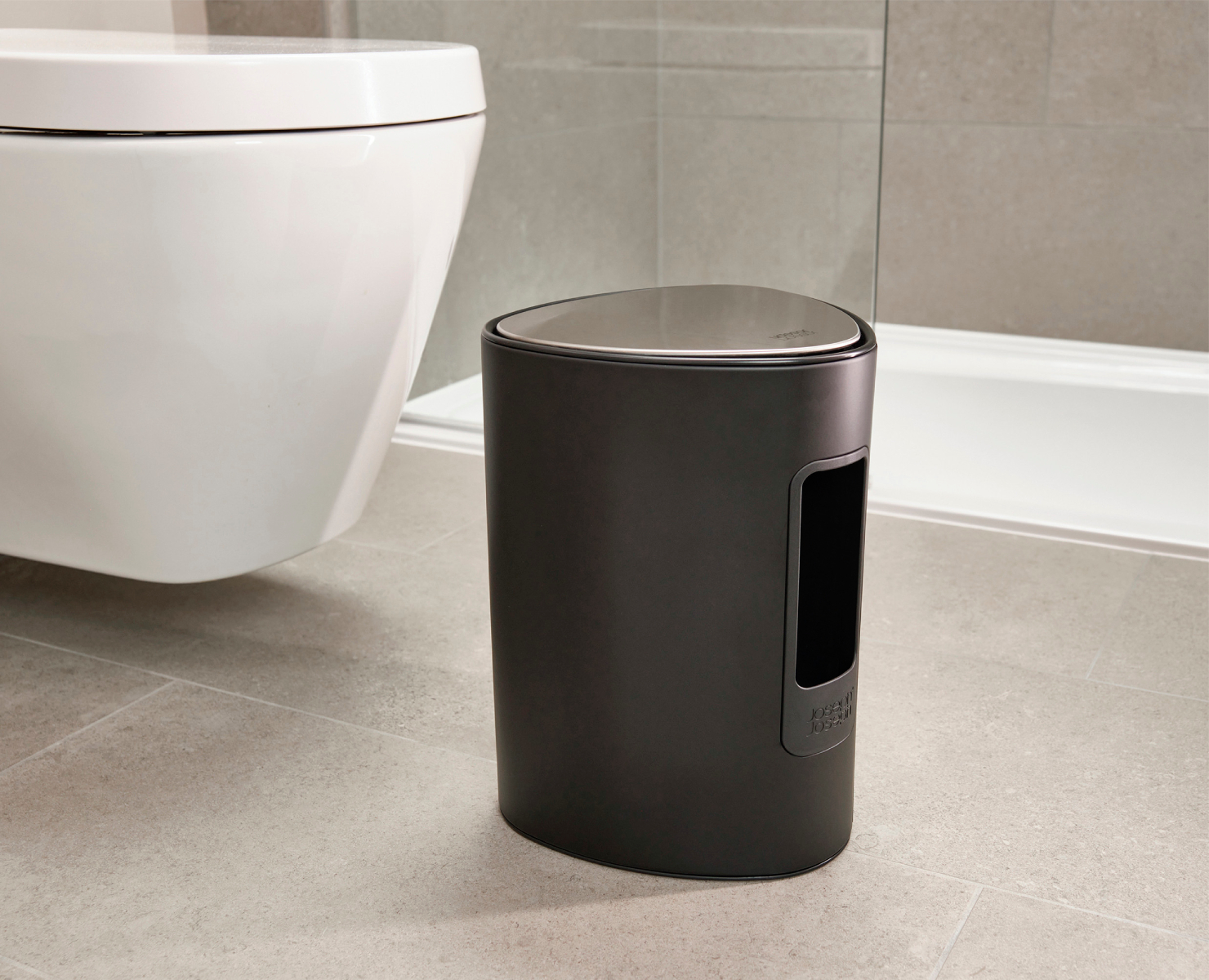 Una escena de baño moderno con un cubo de basura negro, un inodoro blanco y un suelo de baldosas. Una ducha es visible en el fondo.