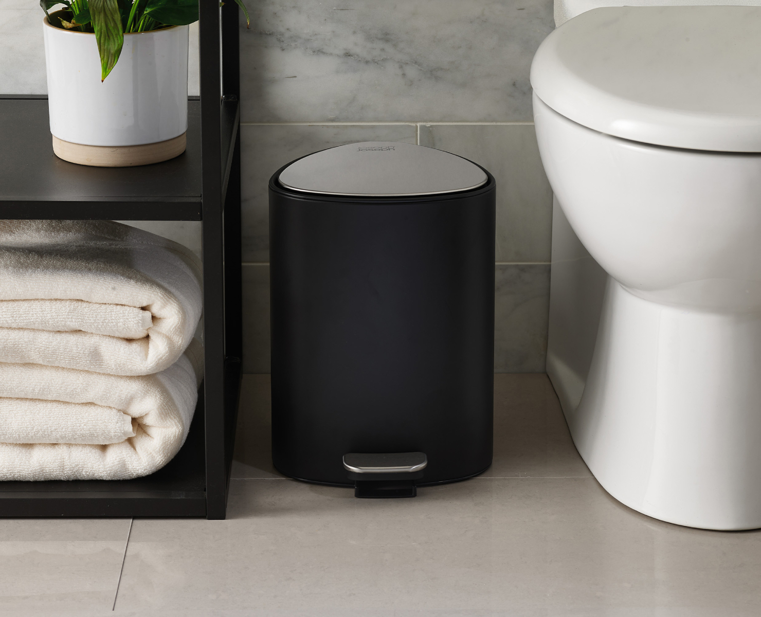 Escena de baño con papelera negra, inodoro blanco y estantería con toallas y planta en maceta.