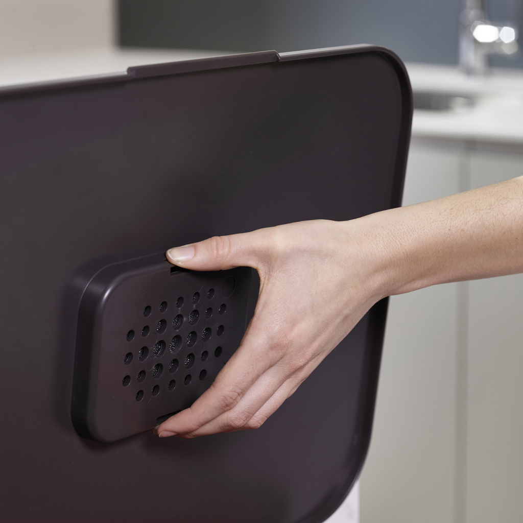 Una mano tiene in mano un oggetto grigio scuro con sezioni perforate. L'oggetto è contro una superficie grigio scuro simile. Una cucina sfocata è sullo sfondo.