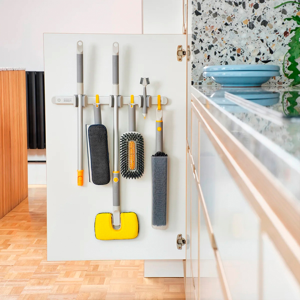 Herramientas de limpieza colgando dentro de una puerta de armario blanco: una fregona, cepillos y un plumero. Una encimera de cocina está a la derecha.