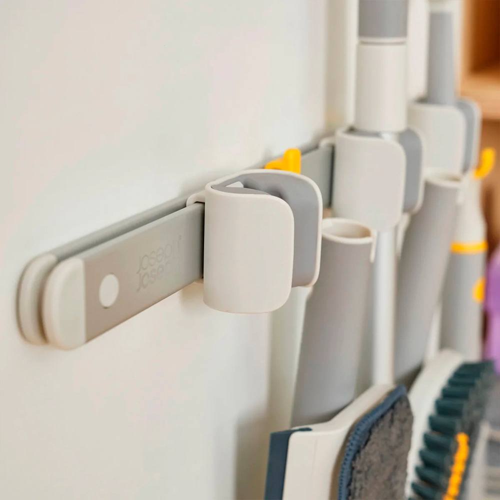 Riel de almacenamiento gris montado en la pared con soportes blancos. Se insertan herramientas de limpieza. La pared es blanca.