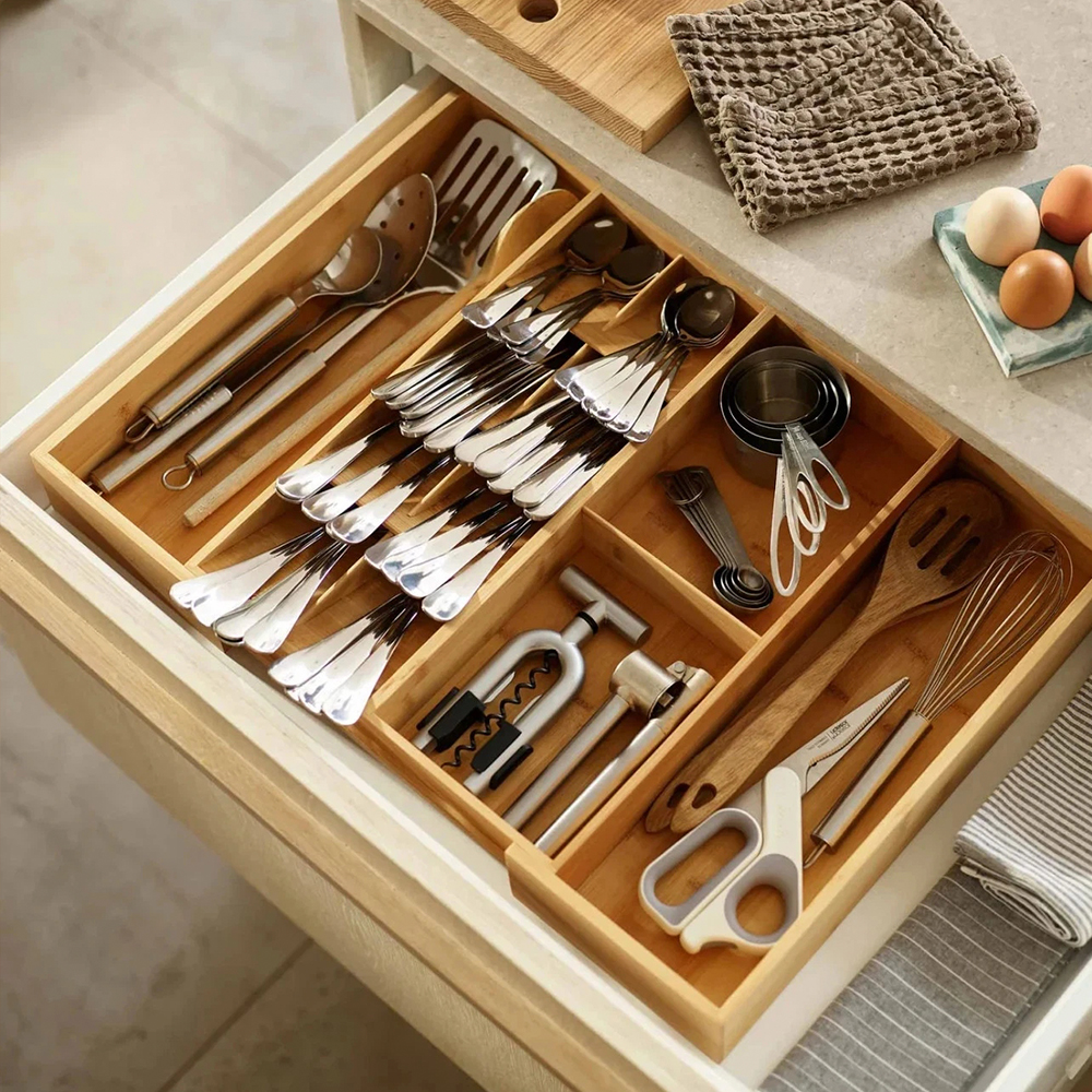 Un cajón de cocina abierto lleno de cubiertos, utensilios y herramientas. Huevos, un paño de cocina y una tabla de cortar de madera están en la encimera.
