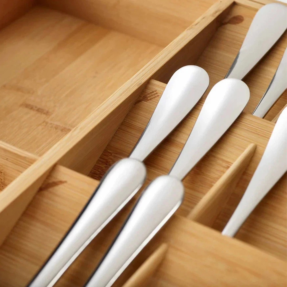 Primer plano de un organizador de cubiertos de madera con varias cucharas plateadas brillantes en su interior, ordenadas. La madera es clara.