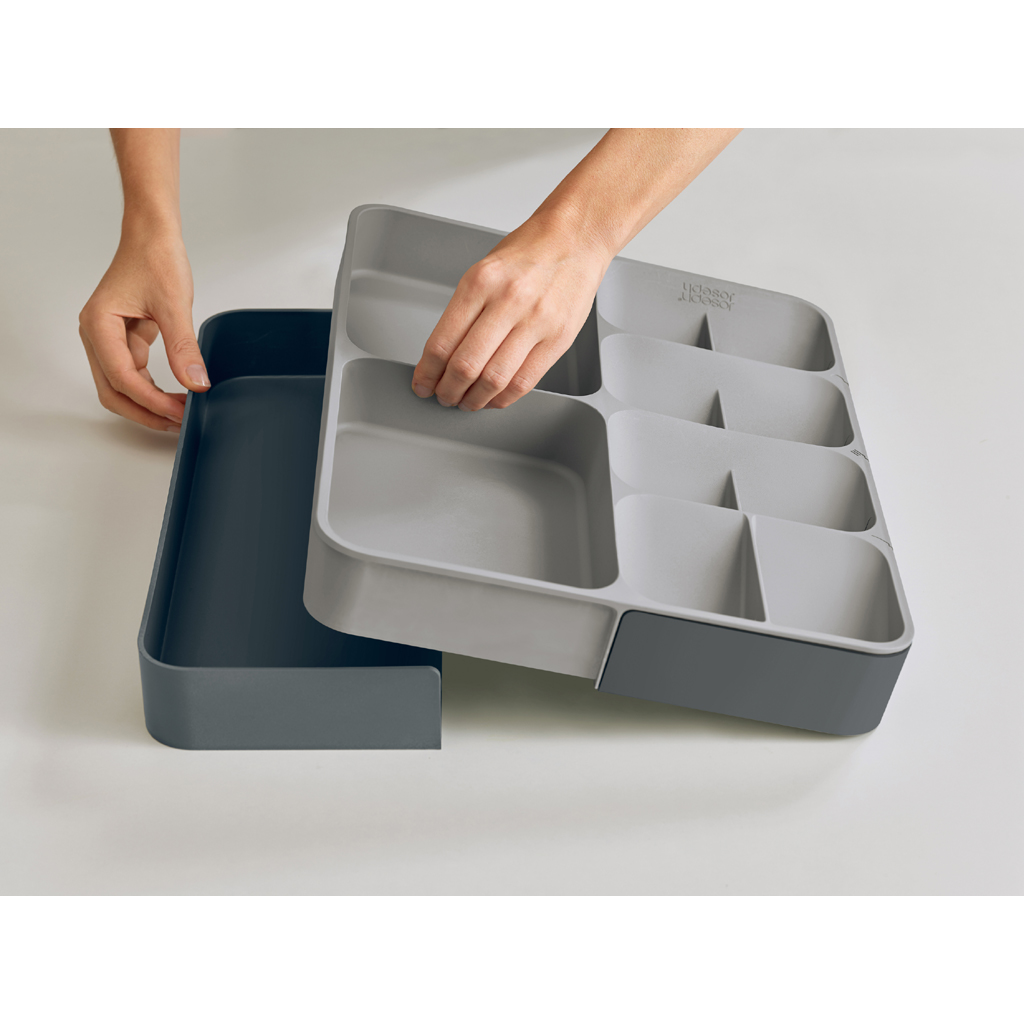 Manos sostienen un organizador de cajones gris. Tiene compartimentos. Debajo, una bandeja gris oscuro está parcialmente extraída sobre una superficie blanca.