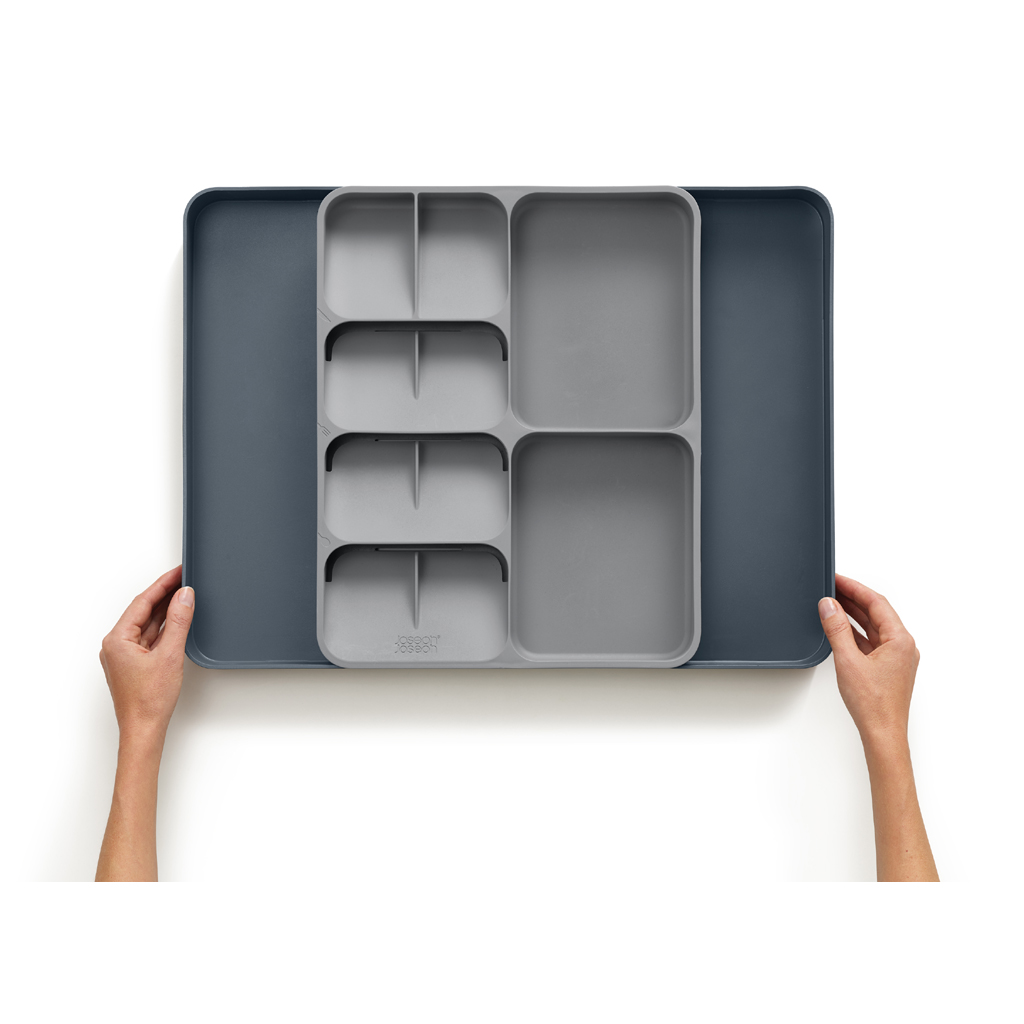 Un organizador de cajones gris con múltiples compartimentos se encuentra dentro de una bandeja gris oscuro más grande. Manos sostienen la bandeja sobre fondo blanco.