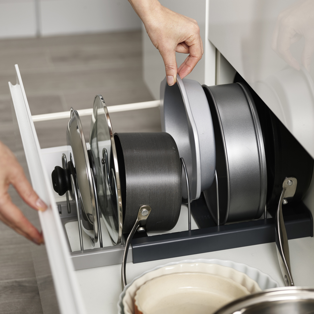 Eine Hand greift in eine Schublade voller Kochgeschirr. Töpfe, Pfannen und Deckel sind ordentlich in einem Gestell verstaut. Die Schublade ist geöffnet.