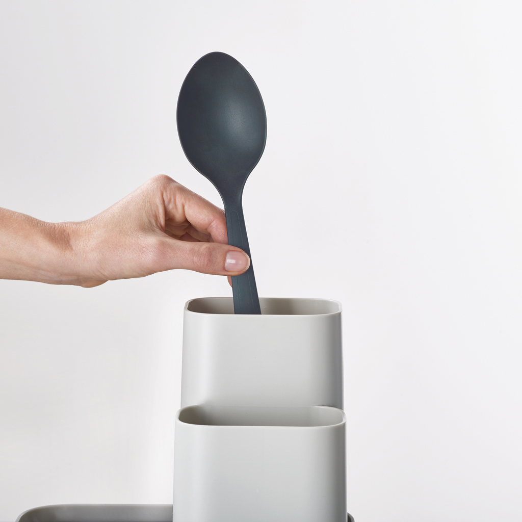 Una mano sostiene una cuchara gris oscuro, colocándola en un soporte para utensilios gris claro. El fondo es blanco.