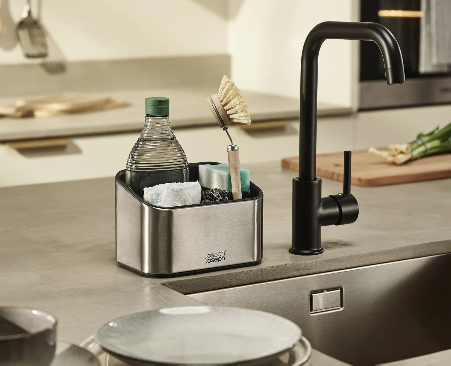 Un fregadero de cocina con un grifo negro. Un organizador de acero inoxidable contiene jabón, esponjas y un cepillo. Una botella de jabón dentro.
