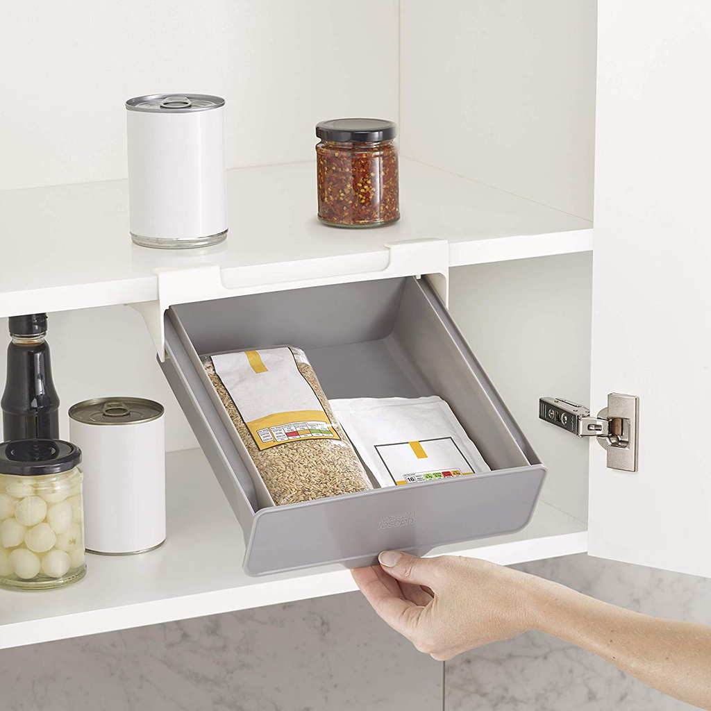 Un armario de despensa con un cajón extraíble gris que contiene dos paquetes de alimentos. Latas y frascos en estantes. Una mano tira del cajón.