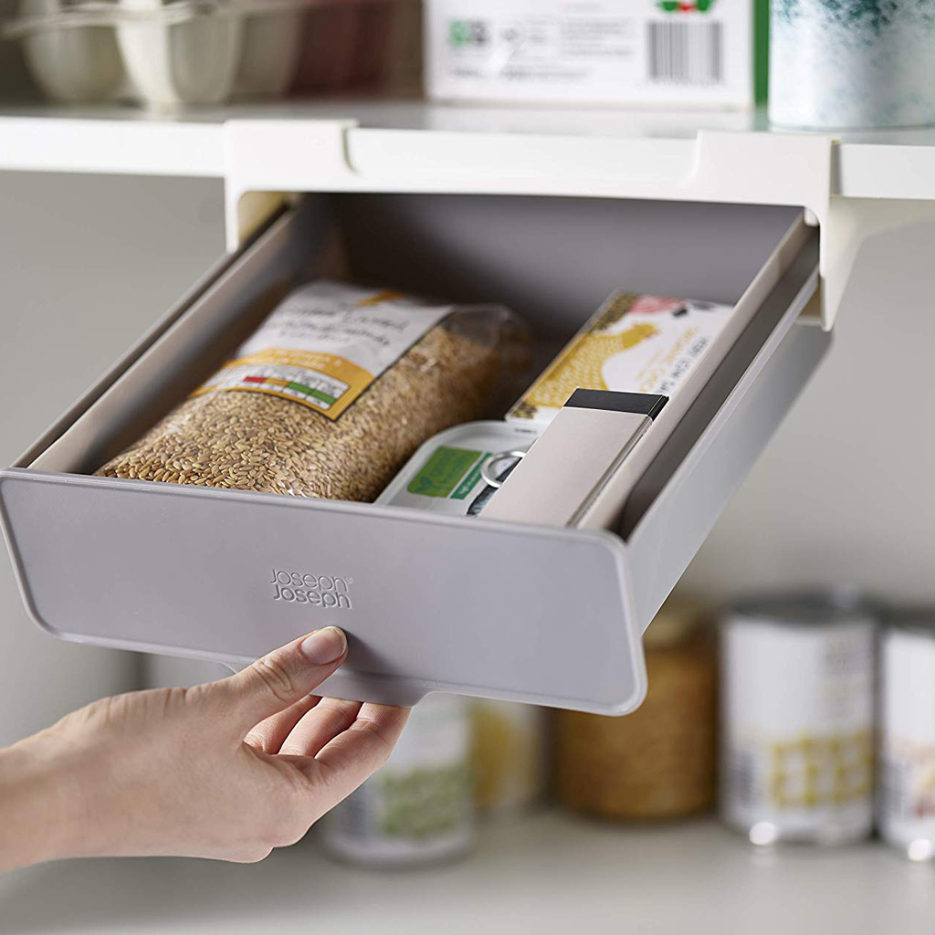 Una mano saca un cajón gris debajo del estante lleno de paquetes de alimentos. El fondo tiene estantes con varios artículos.