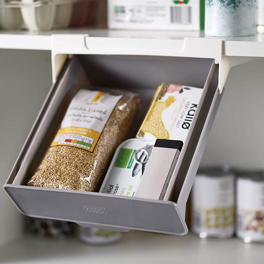 Un cajón gris extraíble debajo de un estante está lleno de alimentos: una bolsa de granos, una caja de pollo y una lata de pescado.