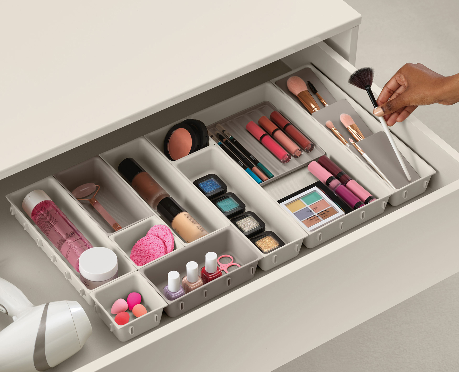 Cajón abierto lleno de maquillaje. Los artículos incluyen brochas, sombras de ojos, labiales, esmalte de uñas y base. Una mano sostiene una brocha de maquillaje.