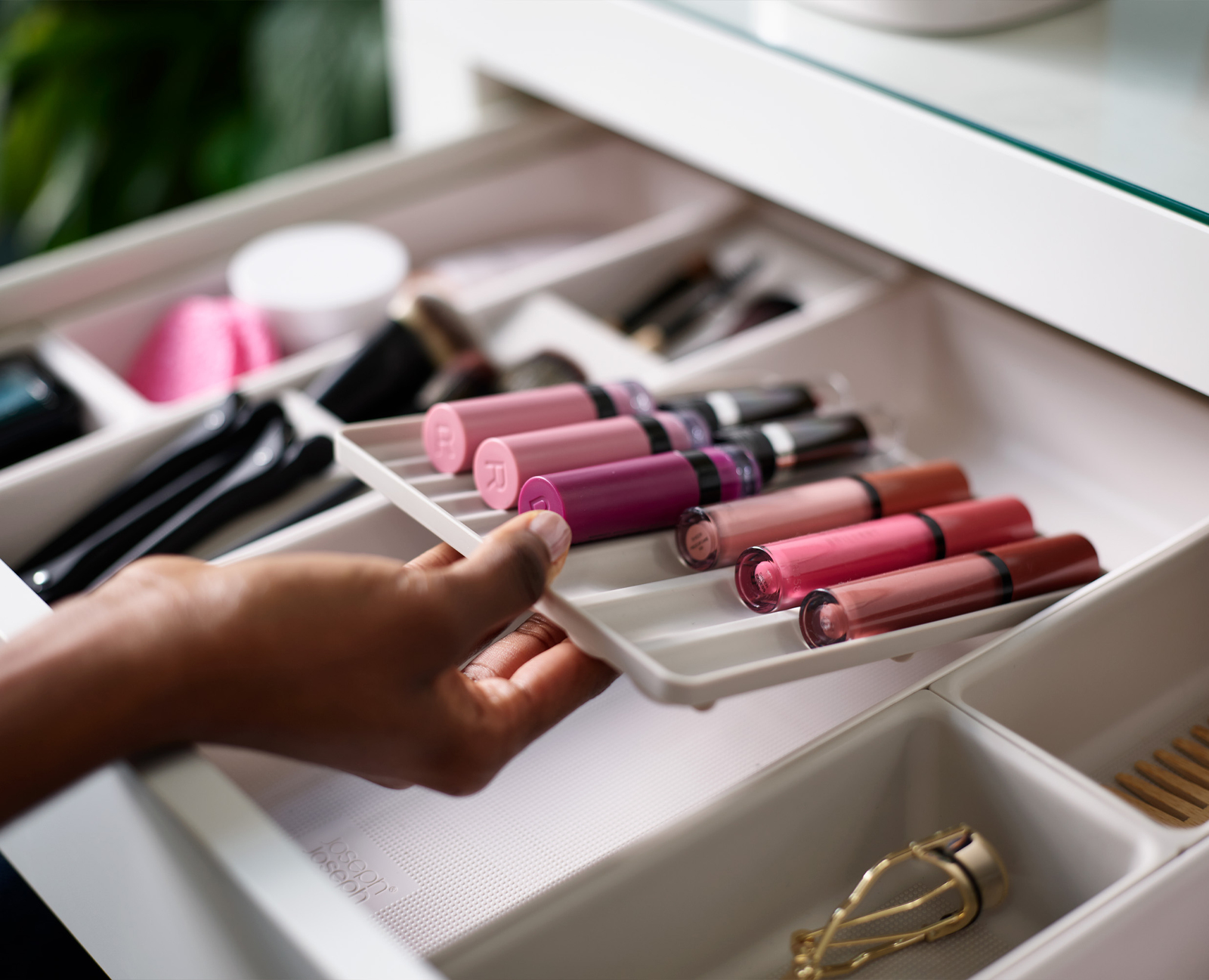 Una mano sostiene una bandeja de brillo labial en un cajón. Varios artículos de maquillaje rodean los brillos.
