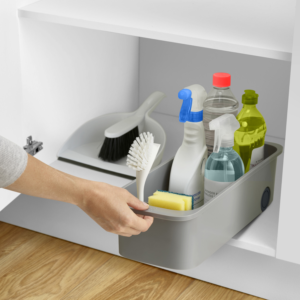 Una mano saca un organizador gris de productos de limpieza de un armario blanco. Contiene botellas, cepillos y una esponja.