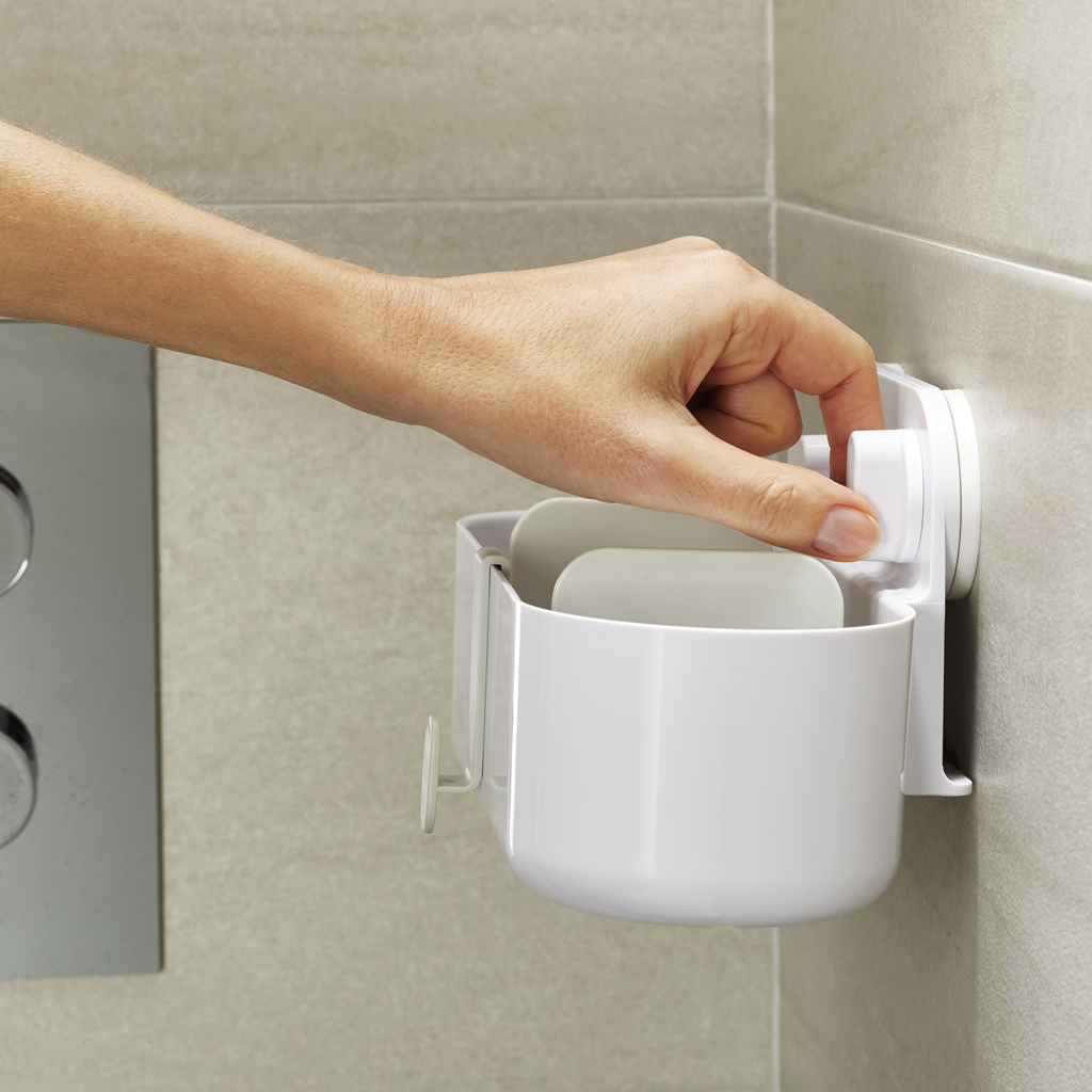 Una mano ajusta un organizador de baño de plástico blanco montado en una pared de azulejos. El organizador contiene artículos y tiene una ventosa.