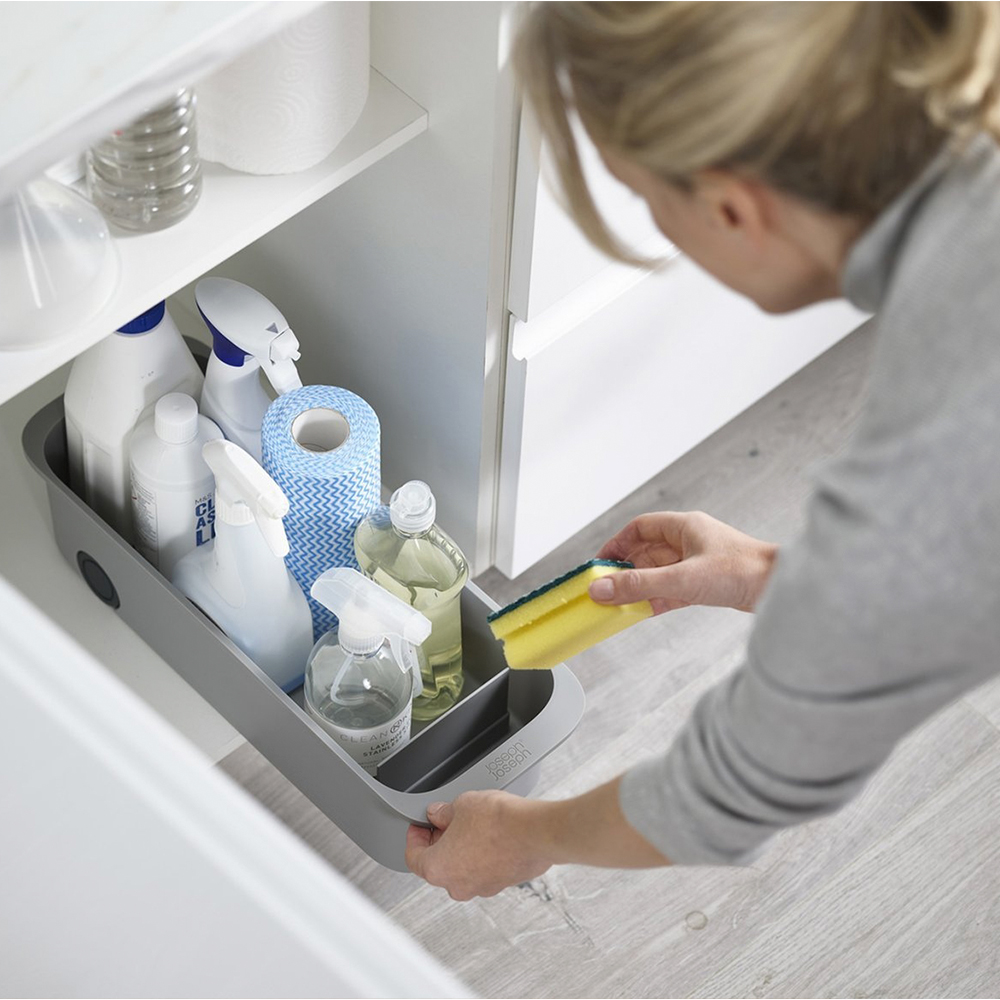 Una mujer sostiene una esponja amarilla, buscando productos de limpieza en un contenedor gris dentro de un armario blanco.